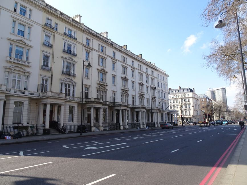A wide view of a multi-storey white Victorian-style residential building situated along a city street, with ornate architectural details including decorative cornices, window moldings, and wrought-iron balconies. The building's façade is clean and well-maintained, with tall, evenly spaced sash windows. In front of the building, a paved sidewalk runs parallel to the road, which has multiple lanes marked with white dashed lines and a solid red line on the right edge. Several parked cars are lined up along the curb, and a few pedestrians are visible walking along the sidewalk. A tall streetlamp extends from the sidewalk near the right side of the image, with additional streetlights further down the street. On the right, a tree with bare branches suggests early spring or late autumn, casting shadows across the pavement. The background features modern high-rise buildings under a bright blue sky with a few scattered clouds. The scene is an urban environment where rubbish removal services, like those offered by Rubbish Clearance Bloomsbury, may operate to support maintaining street cleanliness and private waste clearance.