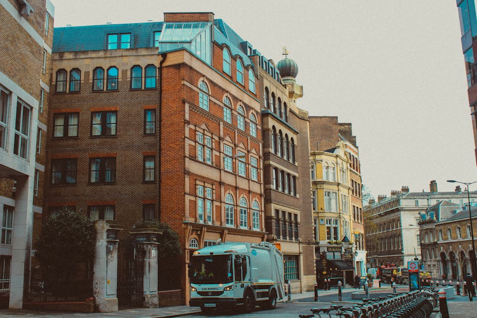 A street scene in an urban area showing a row of historic multi-storey buildings with varied architectural styles, including red brick facades and ornate window frames, alongside a yellow building with decorative detailing. The street features a white waste collection truck parked near a sidewalk with black bollards, and the vehicle is close to an alleyway entrance bordered by stone pillars and greenery. The scene is illuminated by natural daylight, with surrounding infrastructure such as lampposts and bicycles visible, highlighting busy city life. The presence of the waste collection truck and the urban environment suggest services related to rubbish removal or private waste handling, aligning with practices of independent rubbish clearance in the cityscape. The background includes additional historic and modern building facades, with the overall atmosphere emphasizing urban waste management activities and city infrastructure supporting rubbish collection services, as provided by companies like Rubbish Clearance Bloomsbury.