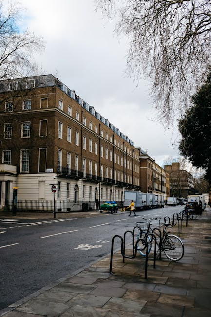 A wide view of a multi-storey white Victorian-style residential building situated along a city street, with ornate architectural details including decorative cornices, window moldings, and wrought-iron balconies. The building's façade is clean and well-maintained, with tall, evenly spaced sash windows. In front of the building, a paved sidewalk runs parallel to the road, which has multiple lanes marked with white dashed lines and a solid red line on the right edge. Several parked cars are lined up along the curb, and a few pedestrians are visible walking along the sidewalk. A tall streetlamp extends from the sidewalk near the right side of the image, with additional streetlights further down the street. On the right, a tree with bare branches suggests early spring or late autumn, casting shadows across the pavement. The background features modern high-rise buildings under a bright blue sky with a few scattered clouds. The scene is an urban environment where rubbish removal services, like those offered by Rubbish Clearance Bloomsbury, may operate to support maintaining street cleanliness and private waste clearance.