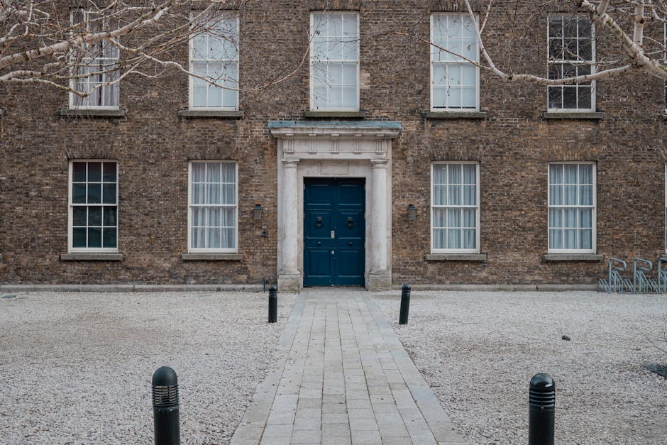 The image depicts the facade of a traditional three-storey brick building with a centrally located dark blue wooden door framed by a white stone surround, featuring decorative molding and a pediment. Six tall, rectangular sash windows with white frames and multiple small panes are symmetrically arranged on the brown brick exterior, with three on each side of the door, some with partially drawn white curtains. The foreground shows a paved pathway made of rectangular stone tiles leading directly to the entrance, flanked by short black bollards on either side, separating the pathway from a surrounding gravel yard composed of light grey, fine gravel. Bare tree branches extend into the upper part of the scene, indicating it is either late autumn or winter. The background consists of the brick wall with some patches of weathering and minimal environmental features, reflecting a quiet, urban residential setting likely adjacent to private property. Rubbish Clearance Bloomsbury offers services that facilitate on-site waste collection, as represented by the organized, unobstructed layout prepared for efficient rubbish removal or clearance activity, although no rubbish is currently visible in the scene.