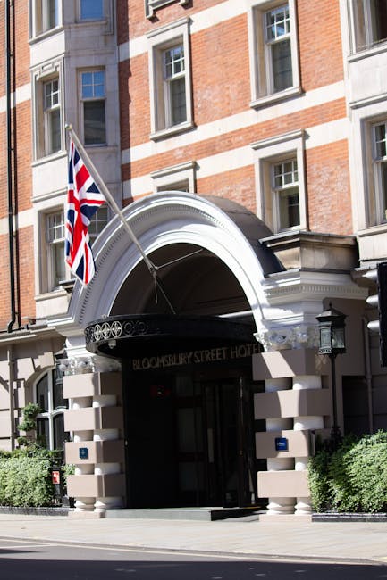 The image displays the entrance to the Bloomsbury Street Hotel, a historic building constructed with red brick and white stone accents, featuring large rectangular windows with white frames. A prominent white archway crowns the doorway, with a black canopy beneath it displaying the hotel’s name in gold lettering. To the left of the arch, a flagpole extends outward, bearing the Union Jack flag, indicating British national identity. Flanking the entrance on either side are neatly trimmed green bushes, adding a touch of greenery to the urban setting. The sidewalk in front is clear, with a smooth paving surface, and the overall scene is illuminated by natural daylight, casting soft shadows. This building’s appearance, characterized by traditional architectural details, suggests an older, well-maintained structure that may be subject to private ownership or independent management, aligning with alternative waste handling or rubbish collection options that could serve buildings of this type or surrounding properties. Rubbish Clearance Bloomsbury often provides on-site clearance services for such urban establishments, ensuring proper waste management according to local standards. The scene emphasizes the building’s exterior detail and urban context, relevant to discussions of private rubbish removal in central London areas.